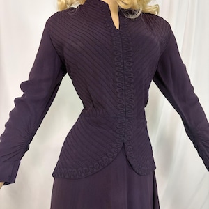 May include: A dark purple long-sleeved blouse featuring a high neckline with a small slit, diagonal pleated panels on the chest, and decorative braided trim running vertically along the center front and around the hem of the bodice.