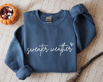 May include: A blue sweatshirt with the words "sweater weather" in white script and a small heart. The sweatshirt is a solid color and has a crew neck. A small orange pumpkin-shaped bowl filled with candy is in the background.