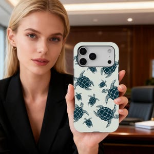 May include: A phone case with a sea turtle pattern. The case is off-white with a repeating pattern of dark blue sea turtles. The phone case has a camera cutout and is held by a person.