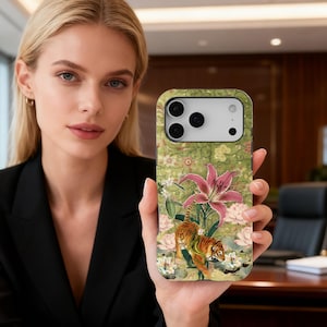 May include: A phone case featuring a floral and tiger design. The case has a green background with pink lilies, white flowers, and two orange and brown tigers. The phone case is held by a person.