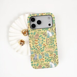 May include: A yellow phone case featuring a pattern of rabbits, flowers, and foliage. The case has a camera cutout and a protective bumper. Two gold-coloured earrings are next to the phone case on a white, ruffled surface.