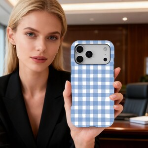 Puede incluir: Funda de teléfono con estampado de cuadros vichy azul claro y blanco. La funda tiene forma rectangular con esquinas redondeadas y un recorte para la cámara. La funda se sostiene frente a una persona.