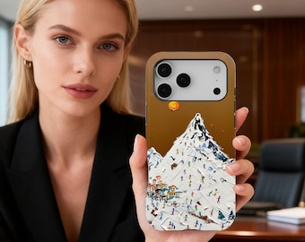 May include: A gold-colored phone case featuring a mountain scene with skiers and a small village. The artwork depicts a snow-covered mountain with a bright orange sun. The case is held by a person.