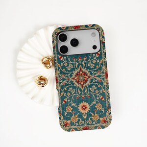 May include: A rectangular smartphone case featuring a teal background with a repeating floral and vine pattern in shades of red, cream, and gold. The case has a cutout for a triple-lens camera system and is positioned next to two gold rings.