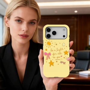 May include: A yellow phone case with a design featuring a sun, stars, a pink bow, and the text "be a light for all to see - Matthew 5:16". The case is held by a person.