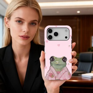 Puede incluir: Funda de teléfono rosa con un dibujo de una rana con un kimono rosa con detalles de fresas. La funda tiene un fondo rosa claro con pequeños corazones y fresas rosas. La funda tiene un recorte para la cámara.