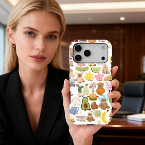Pode incluir: Uma capa de telefone branca com um padrão repetido de ilustrações de desenhos animados fofos. O design inclui ursinhos de pelúcia, abacates, bolos e outros gráficos de alimentos e animais. A capa é segurada por uma pessoa.