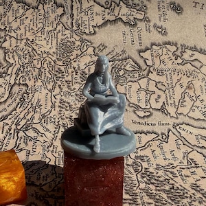 May include: A grey 3D printed miniature of a woman sitting on a throne, holding a book. The miniature is on a grey square base, which is on a red and grey surface. The background is a vintage map.