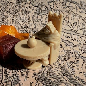 May include: A miniature figurine of a robed figure sitting on a throne with a round table in front of them. The figure is holding a small orb in their hand. The figurine is made of a light brown material and is set against a background of a map.