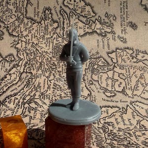 May include: A gray plastic miniature figurine of a person holding a sword. The figurine is standing on a gray plastic base. The figurine is on a brown surface with a map printed on it.