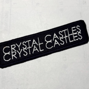 以下が含まれることがあります： 黒い長方形のパッチ。白い糸で「CRYSTAL CASTLES」の文字が刺繍されています。テキストは重ねて3回繰り返されています。パッチには黒い縁取りがあり、アイロン接着または縫い付けのパッチである可能性があります。