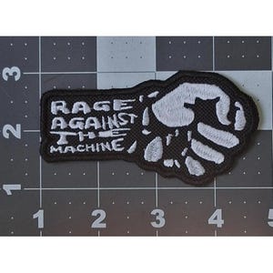 May include: A black and white embroidered patch featuring a clenched fist and the text RAGE AGAINST THE MACHINE. The patch is positioned on a cutting mat with visible inch markings, showing it measures approximately 4 inches in width.