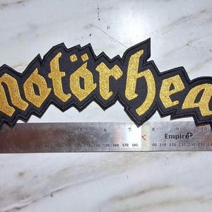 May include: A black and gold embroidered patch with the word "Motörhead" in a gothic font.
