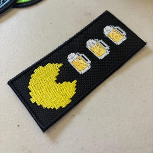 May include: Black rectangular patch featuring a yellow pixelated Pac-Man character on the left and three pixelated beer mugs on the right. The patch is likely an embroidered or woven design.