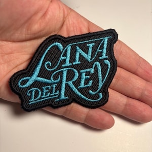 Op de afbeelding: Een zwart geborduurd embleem met de woorden "LANA DEL REY" in turquoise script. De patch heeft een zwarte rand en wordt in een hand gehouden.