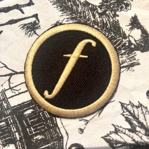 May include: A round, black patch with a gold border and a gold script letter "f" in the center. The patch is on a white fabric with a black and white floral and tree branch pattern.