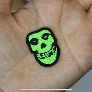 May include: Green and black embroidered iron-on patch of a skull with a toothy grin.