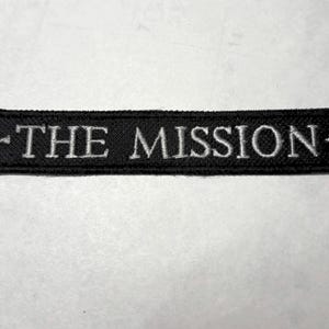 May include: Black rectangular patch with the embroidered text "THE MISSION" in white. The patch has a black border and is likely designed to be sewn onto clothing or accessories. The text is centered.
