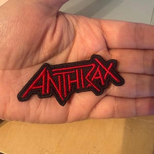 May include: An embroidered patch featuring the word ANTHRAX in a stylized red font with a thick black border. The lettering has a sharp, angular design with a distinct horizontal line extending from the top of the letter A across the word.