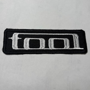 Könnte beinhalten: Schwarzer rechteckiger Aufnäher mit dem Wort "TOOL" in Weiß gestickt. Der Aufnäher hat einen schwarzen Rand und ist zum Aufnähen oder Aufbügeln auf Kleidung oder Accessoires gedacht. Das Design ist ein stilisiertes Logo.