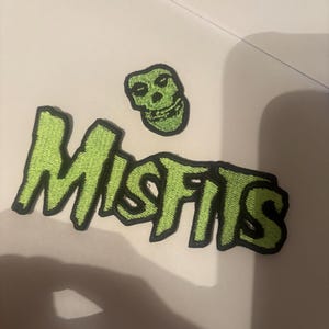 May include: An embroidered patch featuring the word MISFITS in a jagged, lime green font with a black border, accompanied by a separate lime green embroidered skull icon with a black border, both designed for application to fabric surfaces.