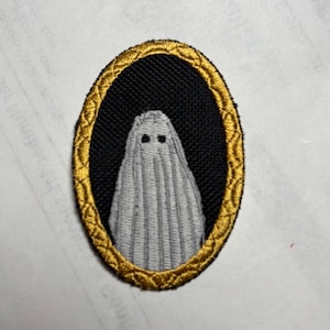 May include: Embroidered patch depicting a white ghost with two small black eyes, on a black background. The ghost is framed by an oval gold border. This patch is designed for application on clothing or accessories.
