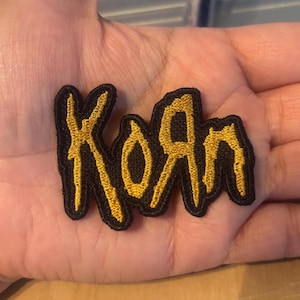 May include: An embroidered patch featuring the band name Korn in a stylized, jagged font. The lettering is stitched in a vibrant gold thread against a solid black background, creating a high-contrast design with a textured, raised appearance.