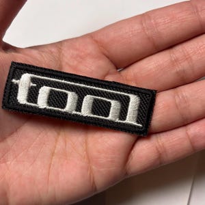 Può includere: Toppa ricamata in bianco e nero con la parola "TOOL" in lettere bianche.