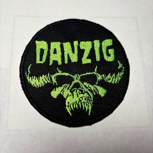 May include: A black and green embroidered patch with the word "DANZIG" in green lettering and a skull with horns in the center.