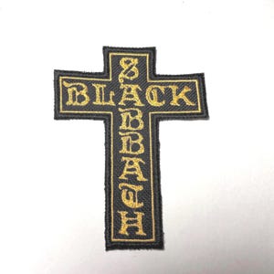 Puede incluir: Un parche bordado en forma de cruz con fondo negro y costuras de color dorado. El texto BLACK SABBATH está bordado en una fuente de estilo gótico, con BLACK en horizontal y SABBATH en vertical a lo largo de la cruz.