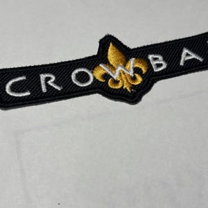 May include: Black embroidered patch with the word "CROW" on the left and "BA" on the right. A gold fleur-de-lis emblem is centered. The patch is on a white surface.