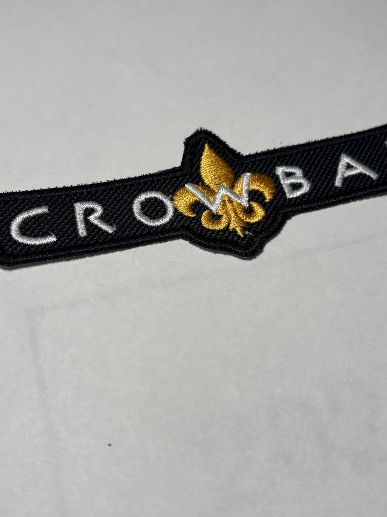 May include: Black embroidered patch with the word "CROW" on the left and "BA" on the right. A gold fleur-de-lis emblem is centered. The patch is on a white surface.