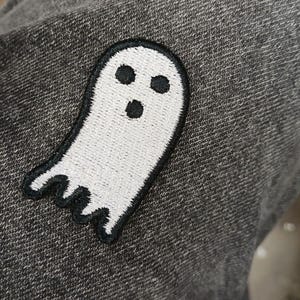 May include: A white ghost patch with a black outline and three black eye and mouth details. The patch is sewn onto a gray fabric, creating a contrast between the bright white ghost and the darker background. The ghost has a friendly, cartoonish appearance.