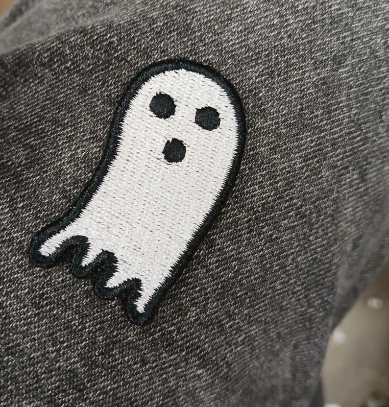 May include: A white ghost patch with a black outline and three black eye and mouth details. The patch is sewn onto a gray fabric, creating a contrast between the bright white ghost and the darker background. The ghost has a friendly, cartoonish appearance.