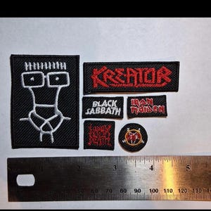 May include: A selection of embroidered patches with various band logos. The patches display the names "Kreator", "Black Sabbath", and "Iron Maiden" in red and white thread on black fabric. A ruler is included for scale, with markings in centimetres.
