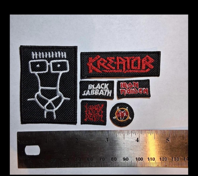 Może przedstawiać: Kolekcja haftowanych naszywek z logo zespoł&oacute;w. Naszywki zawierają nazwy "Kreator", "Black Sabbath" i "Iron Maiden" w czerwonej i białej nici na czarnym materiale. Dołączona jest linijka do skalowania.