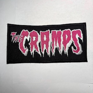 Puede incluir: Parche rectangular negro con el texto "THE CRAMPS" en rosa, contorneado en blanco. El texto tiene un diseño goteante y dentado. El parche está hecho de tela tejida.