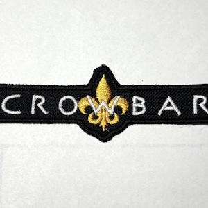 Pode incluir: Patch bordado preto com a palavra "CROWBAR" em letras maiúsculas brancas. Um desenho de flor-de-lis dourada está centrado acima da palavra. O patch é retangular com cantos arredondados.