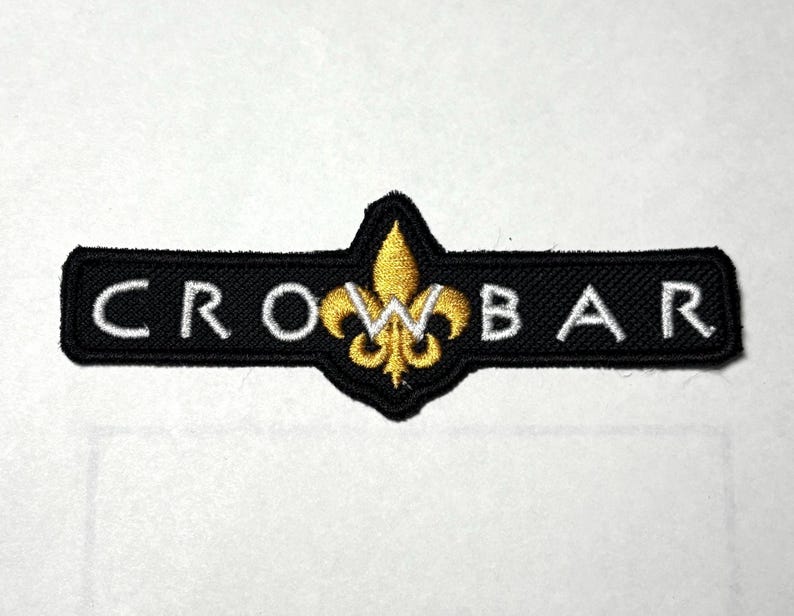 May include: Black embroidered patch with the word "CROWBAR" in white capital letters. A gold fleur-de-lis design is centered above the word. The patch is rectangular with rounded corners.