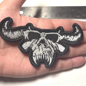 May include: An embroidered patch featuring a stylized silver skull with large, curved horns on a black background. The patch is positioned next to a black ruler, showing a width of approximately 4 inches and a height of approximately 1.5 inches.