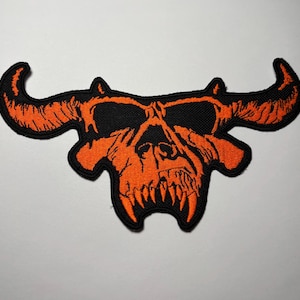 Może przedstawiać: Haftowana naszywka przedstawiająca stylizowaną pomarańczową czaszkę z rogami i ostrymi zębami na czarnym tle. Naszywka ma szczegółowy wzór i jest przeznaczona na odzież lub akcesoria.