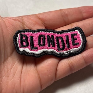 May include: An embroidered patch featuring the word BLONDIE in bold black capital letters. The text is set against a vibrant pink background with a white border, all outlined by a thick black stitched edge, creating a rectangular shape.