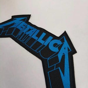 May include: Black and blue embroidered patch with the word "Metallica" in a stylized font.
