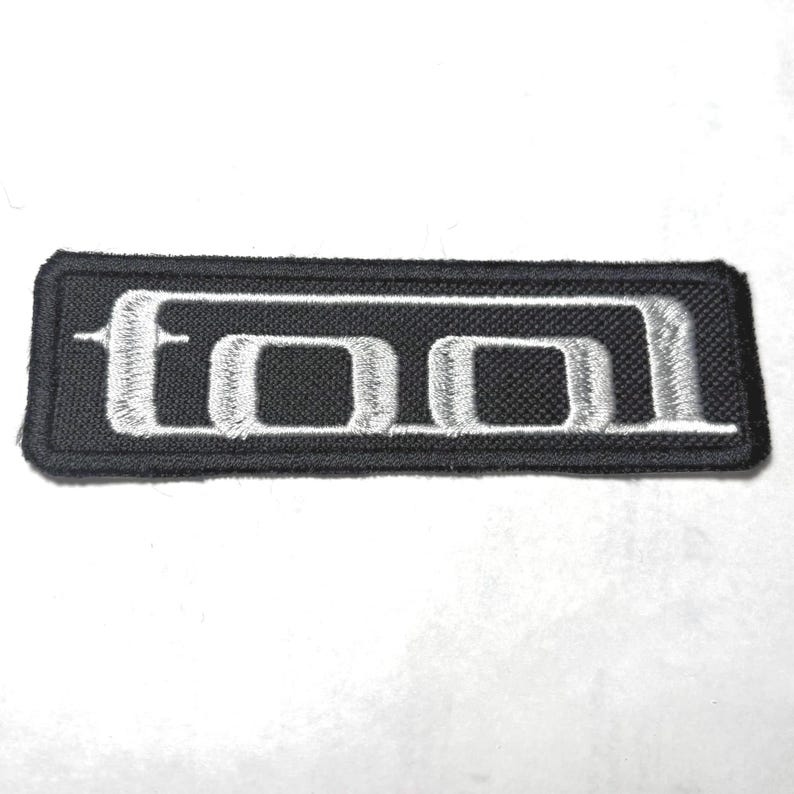 Puede incluir: Un parche rectangular bordado que presenta un fondo negro con la palabra TOOL cosida en hilo blanco. Las letras tienen una fuente estilizada distintiva con una l&iacute;nea horizontal que atraviesa el centro de las letras, creando un aspecto llamativo.