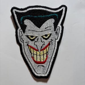Pode incluir: Patch bordado com o rosto do Joker. O patch tem uma borda preta, rosto cinza, olhos amarelos, lábios vermelhos e um sorriso largo revelando dentes amarelos. O cabelo é preto com uma risca verde.