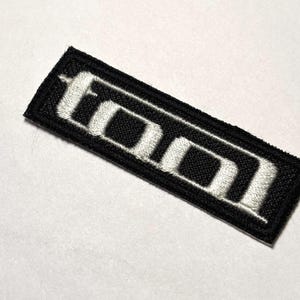 May include: Black fabric patch with white embroidered letters spelling "TOOL" in a stylized font.