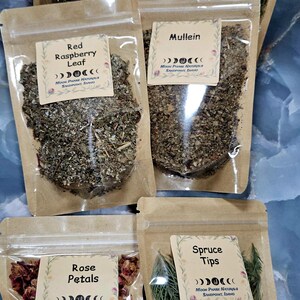 Może przedstawiać: Sześć przezroczystych, zamykanych torebek zawierających suszone zioła i rośliny, każda z kremową etykietą z napisem Moon Phase Naturals, Sandpoint, Idaho. Etykiety określają zawartość jako liść mniszka lekarskiego, igły sosny białej, liść maliny właściwej, dziewannę, płatki r&oacute;ży i pędy świerku.