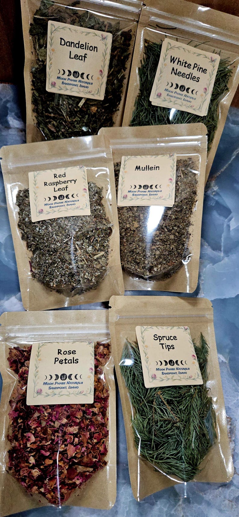 Może przedstawiać: Sześć przezroczystych, zamykanych torebek zawierających suszone zioła i rośliny, każda z kremową etykietą z napisem Moon Phase Naturals, Sandpoint, Idaho. Etykiety określają zawartość jako liść mniszka lekarskiego, igły sosny białej, liść maliny właściwej, dziewannę, płatki r&oacute;ży i pędy świerku.