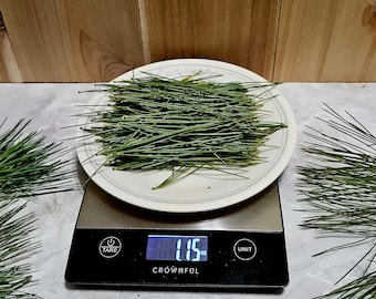 May include: A pile of green pine needles sits on a white plate atop a black digital scale. The scale display shows a weight of 1.15 oz. Additional loose pine needles are scattered on the surface surrounding the scale.