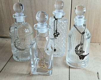 Könnte beinhalten: Vier klare Glasflaschen mit kugelförmigen Verschlüssen, jede mit einer Metallkette und einem Anhänger verziert. Die Anhänger zeigen Sonne und Mond, einen Stern, eine Schlange und einen Schmetterling auf einer Mondsichel.
